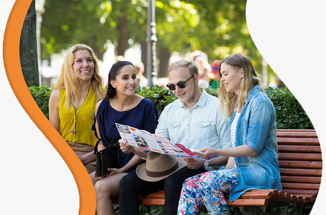 A group of four tourists sitting on a park bench in Helsinki, smiling and reading a Helsinki Card guide. The image represents the ease of exploring the city with the Helsinki Card