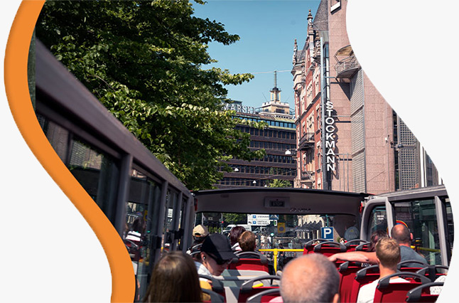 Tourists on a Hop-On Hop-Off double-decker bus tour in Helsinki, passing through the historic city center with red-brick architecture. The bus has an open sunroof, allowing passengers to enjoy panoramic views of the city—one of the many sightseeing experi
